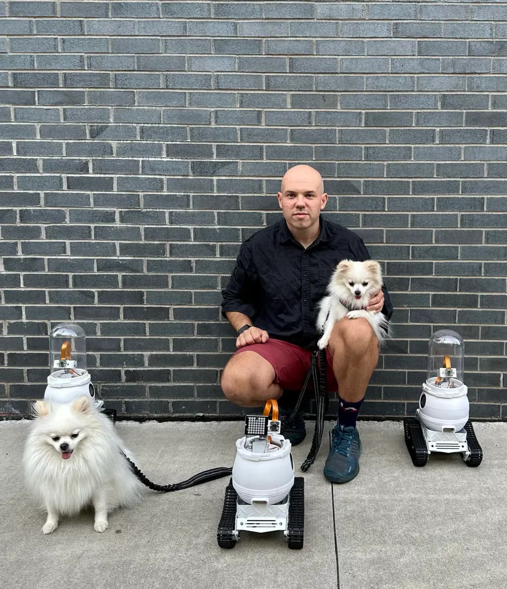 WIM-Z founder kneeling against a brick wall holding a Pomeranian, with prototype rovers beside two dogs